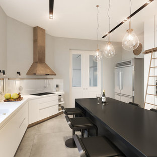 Photo of a contemporary enclosed kitchen in London with a double-bowl sink, flat-panel cabinets, white cabinets, white splashback, glass sheet splashback, stainless steel appliances, concrete flooring, an island, grey floors and white worktops.