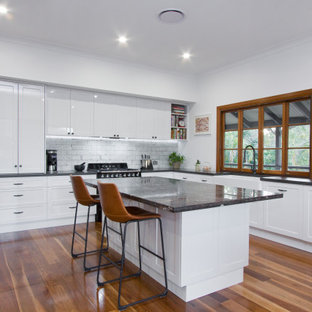 Inspiration for a large transitional l-shaped open plan kitchen in Sunshine Coast with a farmhouse sink, shaker cabinets, white cabinets, granite benchtops, white splashback, subway tile splashback, black appliances, medium hardwood floors, with island and grey benchtop.