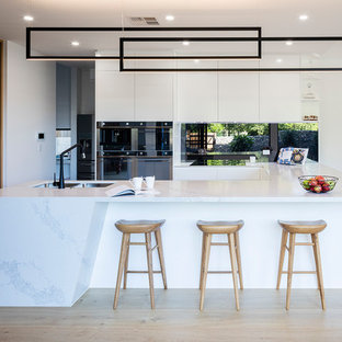 Photo of a mid-sized contemporary u-shaped open plan kitchen in Brisbane with a double-bowl sink, white cabinets, quartzite benchtops, metallic splashback, mirror splashback, black appliances, light hardwood floors, no island, beige floor, white benchtop and flat-panel cabinets.