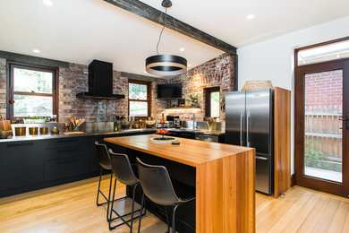 Bild på ett industriellt grå grått l-kök, med en integrerad diskho, släta luckor, svarta skåp, bänkskiva i rostfritt stål, rött stänkskydd, stänkskydd i tegel, rostfria vitvaror, mellanmörkt trägolv, en köksö och brunt golv