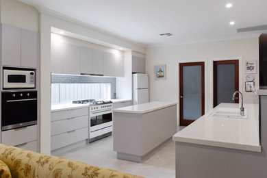 Bild på ett mellanstort funkis vit linjärt vitt kök med öppen planlösning, med en dubbel diskho, släta luckor, grå skåp, bänkskiva i kvarts, stänkskydd med metallisk yta, stänkskydd i glaskakel, vita vitvaror och flera köksöar