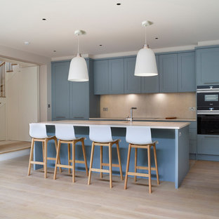 Design ideas for a medium sized nautical kitchen in London with shaker cabinets, blue cabinets, marble worktops, stone slab splashback, an island, white worktops, beige splashback, black appliances, light hardwood flooring and beige floors.