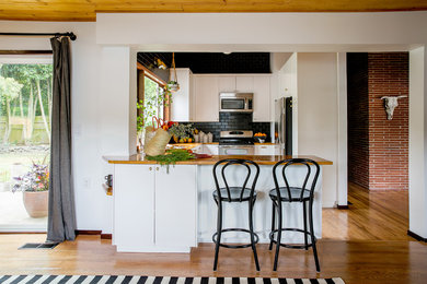 Example of a 1950s u-shaped medium tone wood floor kitchen design in Portland with a peninsula, flat-panel cabinets, white cabinets, wood countertops, black backsplash, subway tile backsplash and stainless steel appliances