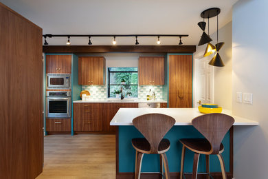 Example of a mid-sized mid-century modern galley medium tone wood floor and brown floor open concept kitchen design in Vancouver with an undermount sink, flat-panel cabinets, medium tone wood cabinets, multicolored backsplash, stainless steel appliances, a peninsula, white countertops and glass tile backsplash