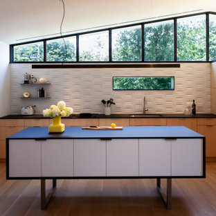 Offene, Mittelgroße Mid-Century Küche in L-Form mit Unterbauwaschbecken, flächenbündigen Schrankfronten, hellen Holzschränken, Laminat-Arbeitsplatte, Küchenrückwand in Weiß, Rückwand aus Keramikfliesen, schwarzen Elektrogeräten, hellem Holzboden, Kücheninsel und schwarzer Arbeitsplatte in New York