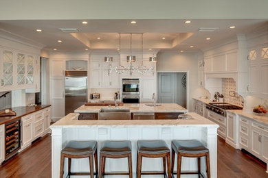 Example of a huge classic u-shaped medium tone wood floor eat-in kitchen design in Jacksonville with a farmhouse sink, beaded inset cabinets, white cabinets, marble countertops, multicolored backsplash, glass tile backsplash, stainless steel appliances and two islands