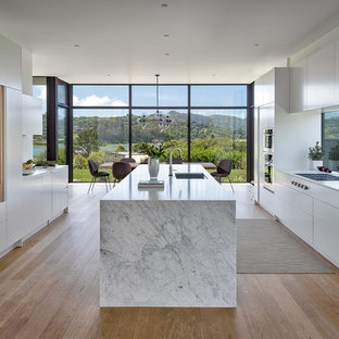 Modelo de cocina comedor minimalista, grande, con fregadero bajoencimera, armarios con paneles lisos, puertas de armario blancas, encimera de mármol, electrodomésticos con paneles, suelo de madera clara, una isla, encimeras blancas, salpicadero de vidrio y suelo beige