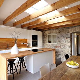 Inspiration for a mid-sized contemporary single-wall eat-in kitchen in Gloucestershire with a double-bowl sink, flat-panel cabinets, white cabinets, wood benchtops, stainless steel appliances, concrete floors and a peninsula.