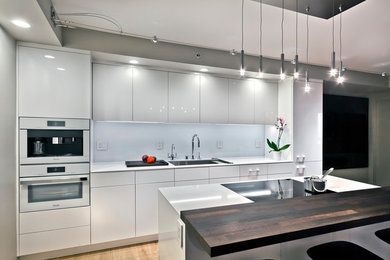 Small single-wall open plan kitchen in Minneapolis with a submerged sink, flat-panel cabinets, white cabinets, engineered stone countertops, white splashback, glass sheet splashback, white appliances, medium hardwood flooring, an island, brown floors and white worktops.