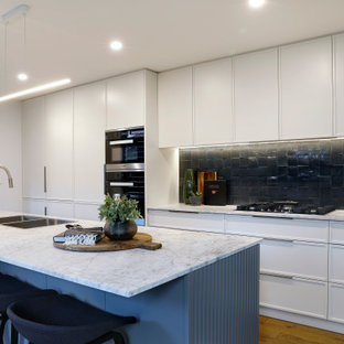 Mid-sized contemporary galley open plan kitchen in Sydney with a drop-in sink, marble benchtops, ceramic splashback, stainless steel appliances, with island, white benchtop, flat-panel cabinets, white cabinets, black splashback, medium hardwood floors and brown floor.