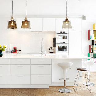 Cette image montre une grand cuisine américaine parallèle design avec un placard à porte plane, des portes de placard blanches, une crédence blanche, un électroménager blanc, un sol en bois clair, un îlot central, un évier posé, un plan de travail en verre recyclé et une crédence en carreau de porcelaine.