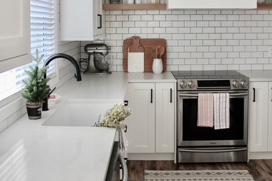Inspiration for a mid-sized farmhouse l-shaped laminate floor and brown floor enclosed kitchen remodel in Vancouver with a farmhouse sink, shaker cabinets, white cabinets, solid surface countertops, white backsplash, ceramic backsplash, stainless steel appliances, no island and white countertops