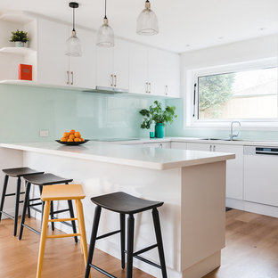 This is an example of a large contemporary u-shaped open plan kitchen in Melbourne with a double-bowl sink, flat-panel cabinets, white cabinets, granite benchtops, green splashback, glass sheet splashback, brown floor, white benchtop, panelled appliances, medium hardwood floors and a peninsula.