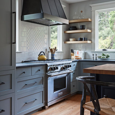 Example of a mid-sized arts and crafts l-shaped light wood floor and beige floor enclosed kitchen design in San Francisco with a farmhouse sink, shaker cabinets, gray cabinets, soapstone countertops, white backsplash, ceramic backsplash, stainless steel appliances and an island