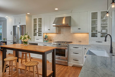 Kitchen pantry - mid-sized country l-shaped light wood floor and brown floor kitchen pantry idea in Boston with a farmhouse sink, flat-panel cabinets, white cabinets, marble countertops, gray backsplash, porcelain backsplash, stainless steel appliances and an island