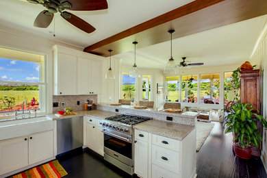 Example of a mid-sized beach style u-shaped bamboo floor and black floor open concept kitchen design in Hawaii with a farmhouse sink, shaker cabinets, white cabinets, granite countertops, gray backsplash, stone slab backsplash, stainless steel appliances and a peninsula