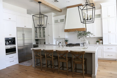 Large trendy l-shaped light wood floor and brown floor kitchen photo with a farmhouse sink, shaker cabinets, white cabinets, stainless steel appliances, an island, white backsplash, ceramic backsplash, quartzite countertops and white countertops