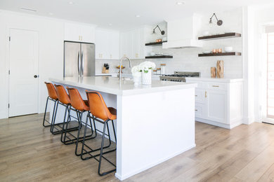 This is an example of a farmhouse kitchen in Orange County with a belfast sink, shaker cabinets, white splashback, metro tiled splashback, stainless steel appliances, light hardwood flooring, an island and white cabinets.