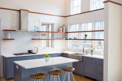 This is an example of a beach style l-shaped open plan kitchen in Boston with a belfast sink, shaker cabinets, blue cabinets, white splashback, marble splashback, medium hardwood flooring, an island, brown floors and stainless steel appliances.