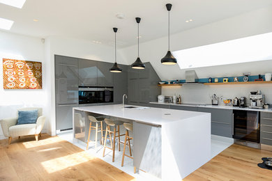 Photo of a large contemporary single-wall kitchen in London with grey cabinets, an island, a submerged sink, flat-panel cabinets, white splashback, integrated appliances, light hardwood flooring and brown floors.