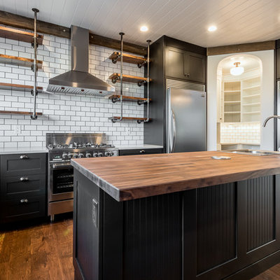 Inspiration for a large industrial u-shaped medium tone wood floor and brown floor kitchen remodel in Chicago with shaker cabinets, black cabinets, wood countertops, white backsplash, subway tile backsplash, stainless steel appliances, an island and a farmhouse sink