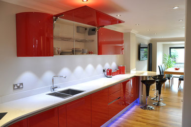 Photo of a large contemporary u-shaped kitchen/diner in Kent with a built-in sink, flat-panel cabinets, red cabinets, quartz worktops, white splashback, stainless steel appliances, medium hardwood flooring, a breakfast bar, brown floors and white worktops.
