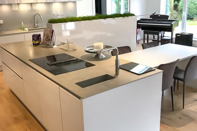 Photo of an expansive modern open plan kitchen in Other with flat-panel cabinets, white cabinets, concrete worktops, glass sheet splashback, black appliances, light hardwood flooring, an island and brown floors.