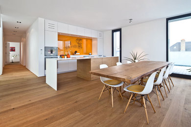 Foto de cocina comedor lineal moderna con suelo de madera en tonos medios, armarios con paneles lisos, puertas de armario blancas, salpicadero naranja, electrodomésticos de acero inoxidable y una isla