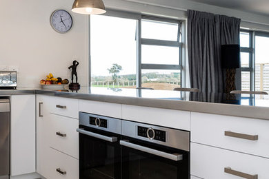 Design ideas for a large modern u-shaped open plan kitchen in Auckland with a double-bowl sink, flat-panel cabinets, white cabinets, stainless steel worktops, white splashback, glass sheet splashback, stainless steel appliances, porcelain flooring, a breakfast bar and grey floors.