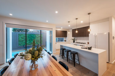 Design ideas for a large contemporary galley eat-in kitchen in Melbourne with flat-panel cabinets, white cabinets, quartz benchtops, white splashback, light hardwood floors, with island, brown floor, white benchtop, an integrated sink, porcelain splashback and white appliances.