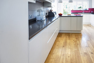 Large modern kitchen in Sussex with a built-in sink, flat-panel cabinets, white cabinets, granite worktops, pink splashback, glass sheet splashback, light hardwood flooring, a breakfast bar and grey worktops.