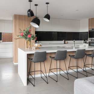 This is an example of a scandinavian l-shaped open plan kitchen in Perth with an undermount sink, flat-panel cabinets, white cabinets, black splashback, glass sheet splashback, stainless steel appliances, with island, grey floor and white benchtop.