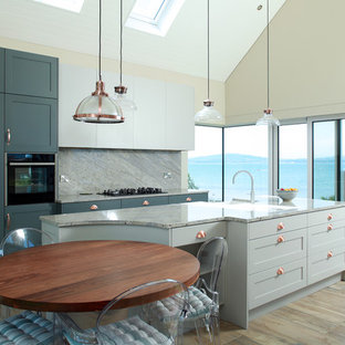 This is an example of a medium sized coastal kitchen in London with shaker cabinets, marble worktops, grey splashback, marble splashback, stainless steel appliances, an island, grey worktops, a belfast sink, blue cabinets, light hardwood flooring and brown floors.