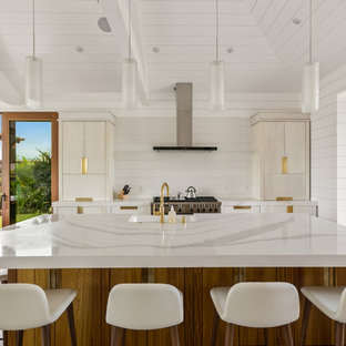 Inspiration for a large beach style galley open plan kitchen in Hawaii with a farmhouse sink, flat-panel cabinets, white cabinets, quartz benchtops, white splashback, stone slab splashback, stainless steel appliances, porcelain floors, with island and grey floor.