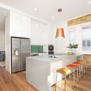 Contemporary single-wall kitchen in Sydney with white cabinets, green splashback, stainless steel appliances, medium hardwood floors, with island, brown floor, grey benchtop, an undermount sink, flat-panel cabinets, solid surface benchtops and mosaic tile splashback.