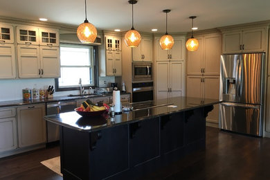 Example of a large farmhouse l-shaped dark wood floor and brown floor eat-in kitchen design in Other with a double-bowl sink, shaker cabinets, beige cabinets, laminate countertops, white backsplash, ceramic backsplash, stainless steel appliances and an island