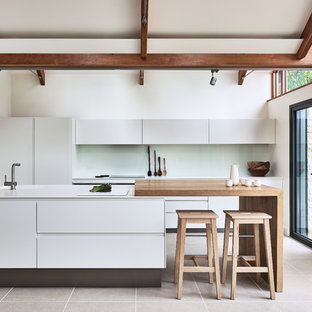 Photo of a large country galley kitchen in Other with an integrated sink, porcelain flooring, beige floors, flat-panel cabinets, white cabinets, green splashback, glass sheet splashback, a breakfast bar and white worktops.