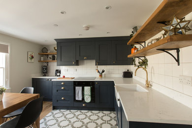 This is an example of a medium sized eclectic l-shaped open plan kitchen in Sussex with a belfast sink, shaker cabinets, black cabinets, composite countertops, white splashback, ceramic splashback, black appliances, vinyl flooring and no island.