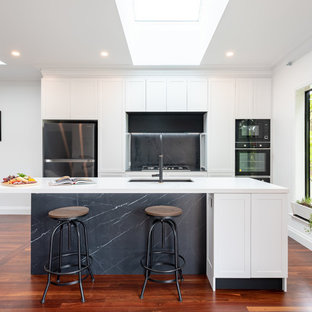 This is an example of a mid-sized transitional galley open plan kitchen in Perth with an undermount sink, shaker cabinets, white cabinets, quartz benchtops, grey splashback, stone slab splashback, black appliances, medium hardwood floors, with island, white benchtop and brown floor.