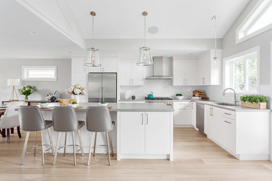 Photo of a medium sized classic l-shaped kitchen/diner in Vancouver with a double-bowl sink, flat-panel cabinets, white cabinets, quartz worktops, white splashback, metro tiled splashback, stainless steel appliances, medium hardwood flooring and an island.