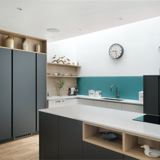 This is an example of a medium sized contemporary l-shaped kitchen/diner in Gloucestershire with flat-panel cabinets, grey cabinets, quartz worktops, blue splashback, glass sheet splashback, black appliances, medium hardwood flooring, white worktops, a submerged sink and a breakfast bar.