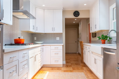 Example of a trendy galley light wood floor, brown floor and vaulted ceiling eat-in kitchen design in San Francisco with a farmhouse sink, shaker cabinets, white cabinets, quartz countertops, gray backsplash, ceramic backsplash, stainless steel appliances, no island and gray countertops