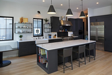 Example of a minimalist light wood floor and brown floor kitchen design in Minneapolis with a farmhouse sink, recessed-panel cabinets, black cabinets, quartz countertops, stainless steel appliances and white countertops