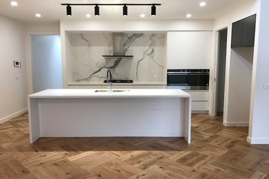 Large modern galley open plan kitchen in Melbourne with a submerged sink, white cabinets, engineered stone countertops, white splashback, porcelain splashback, black appliances, light hardwood flooring and an island.