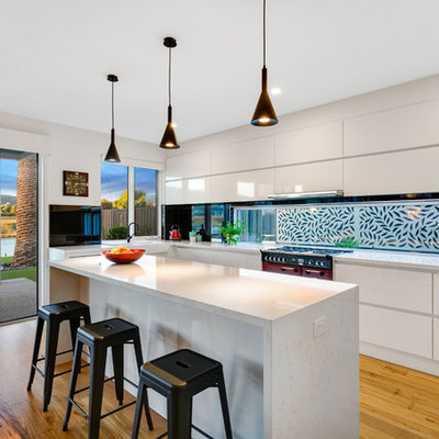 Inspiration for a large contemporary l-shaped light wood floor and beige floor open concept kitchen remodel in Melbourne with a farmhouse sink, flat-panel cabinets, white cabinets, window backsplash, colored appliances, an island, quartz countertops, black backsplash and gray countertops