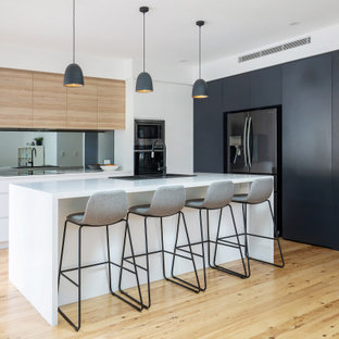 Mid-sized contemporary l-shaped open plan kitchen in Sydney with a drop-in sink, flat-panel cabinets, quartz benchtops, metallic splashback, mirror splashback, light hardwood floors, with island, white benchtop, black appliances and beige floor.