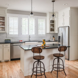 Small transitional open concept kitchen ideas - Example of a small transitional l-shaped light wood floor open concept kitchen design in Nashville with a farmhouse sink, shaker cabinets, white cabinets, white backsplash, subway tile backsplash, stainless steel appliances, an island and black countertops
