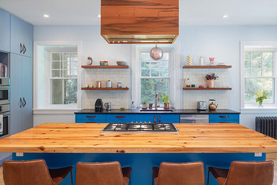 На фото: кухня в современном стиле с врезной мойкой, плоскими фасадами, синими фасадами, деревянной столешницей, белым фартуком, фартуком из плитки кабанчик, техникой из нержавеющей стали и островом