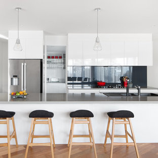 Design ideas for a contemporary galley kitchen in Melbourne with a drop-in sink, flat-panel cabinets, white cabinets, black splashback, glass sheet splashback, stainless steel appliances, medium hardwood floors, with island, brown floor and grey benchtop.