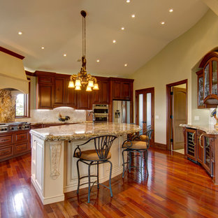 This is an example of an expansive traditional u-shaped open plan kitchen in Denver with an undermount sink, louvered cabinets, medium wood cabinets, granite benchtops, white splashback, limestone splashback, stainless steel appliances, medium hardwood floors, with island and brown floor.